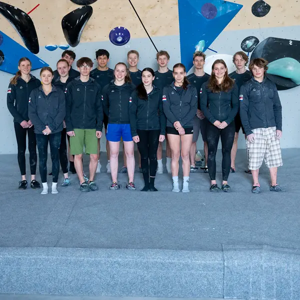 Een groep van 15 jonge sporters in bij elkaar passende jassen staat binnen op een grijs tapijt voor een blauw- en beige klimwand met diverse grepen en volumes. Een groep van 15 jonge sporters in bij elkaar passende jassen staat binnen op een grijs tapijt voor een blauw- en beige klimwand met diverse grepen en volumes.